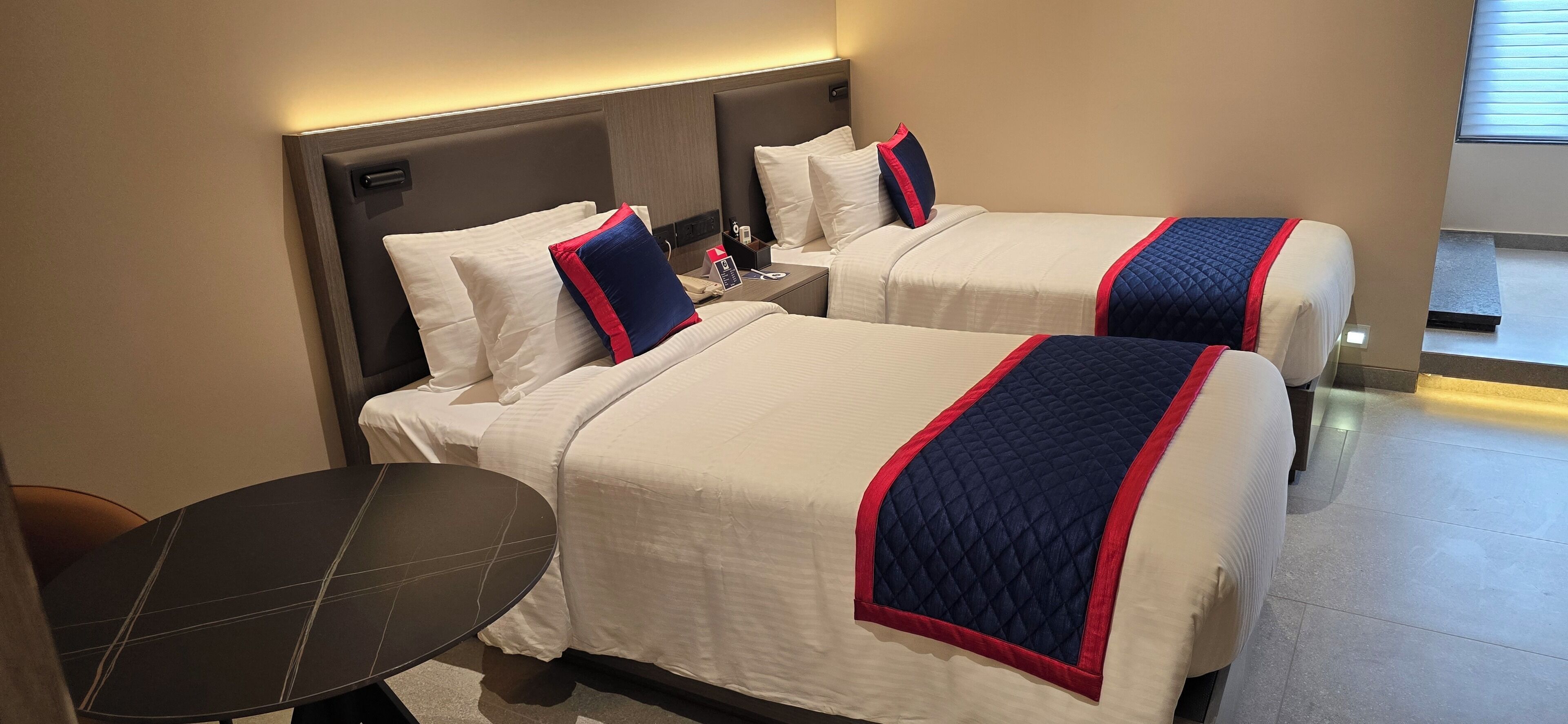 A hotel room with two beds, each with white bedding and blue and red accents. There is a small table and a chair in the corner.