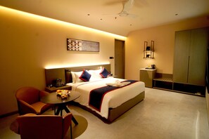 Une chambre d’hôtel comprenant un lit, un coin salon avec une table, une chaise, une petite table avec un saladier de fruits, une table de chevet et une armoire.