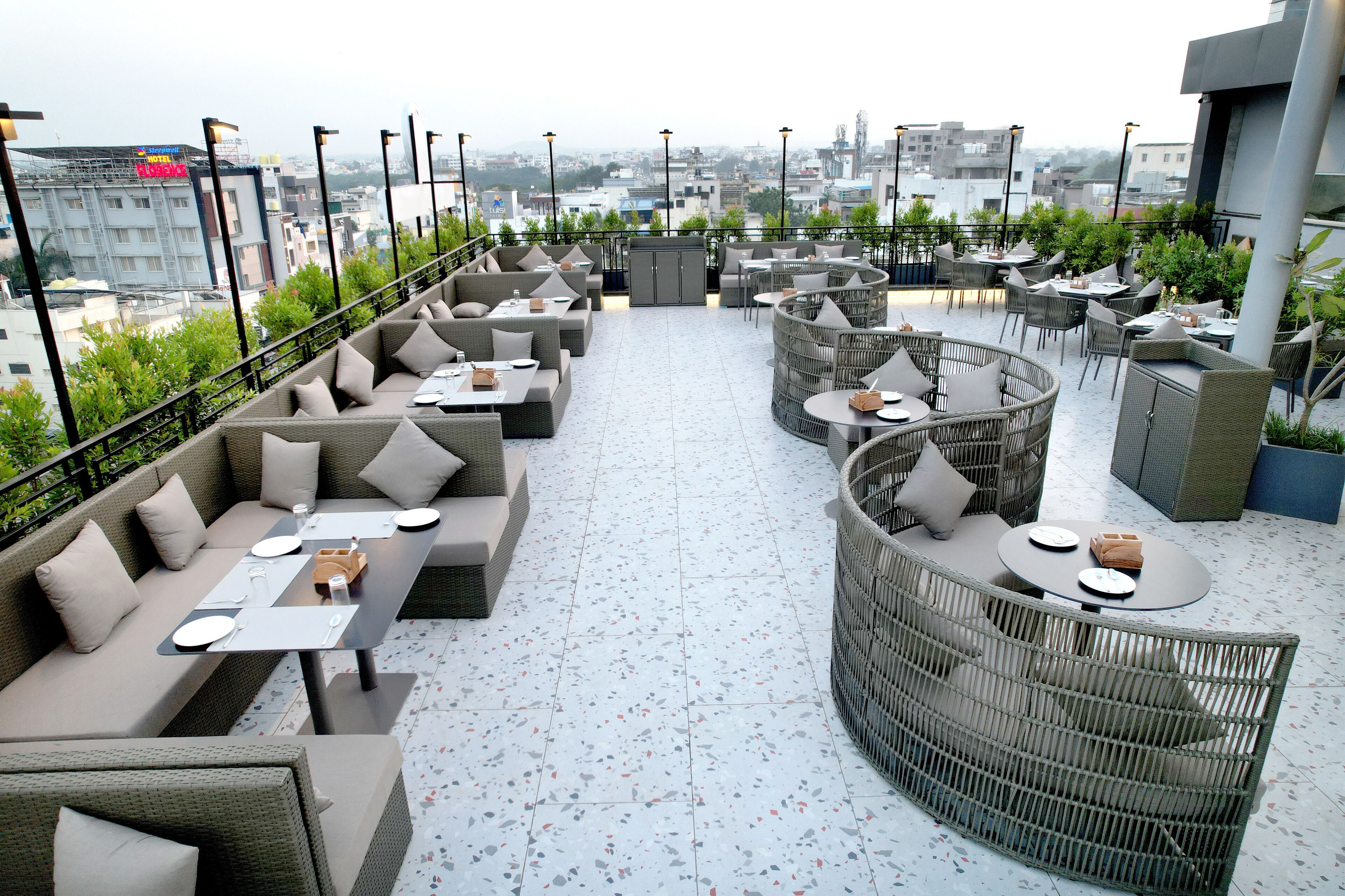 En tagterrasse med udendørs siddepladser, herunder sofaer og runde borde, samt udsigt over byen.