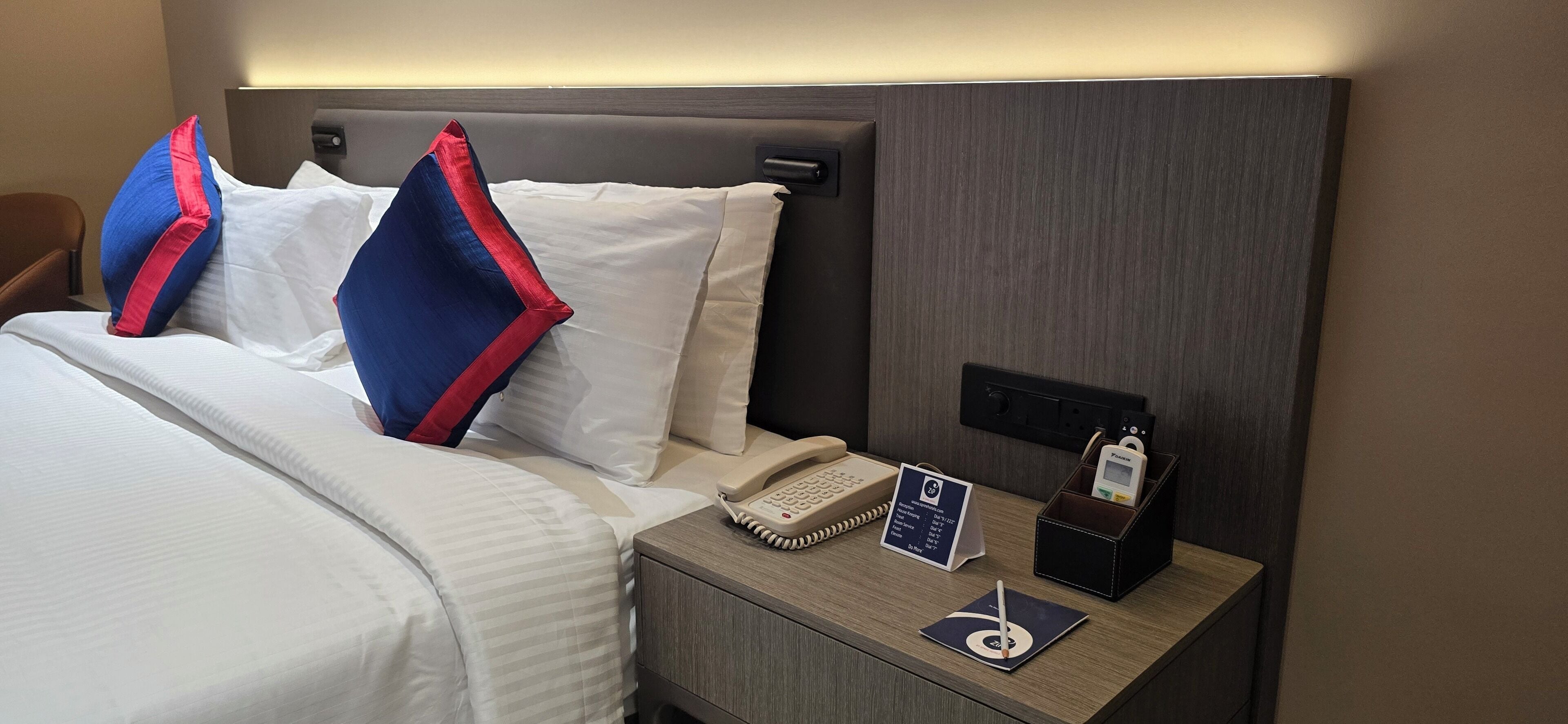 A hotel room with a neatly made bed, a bedside table with a telephone, a notepad, and a pen.