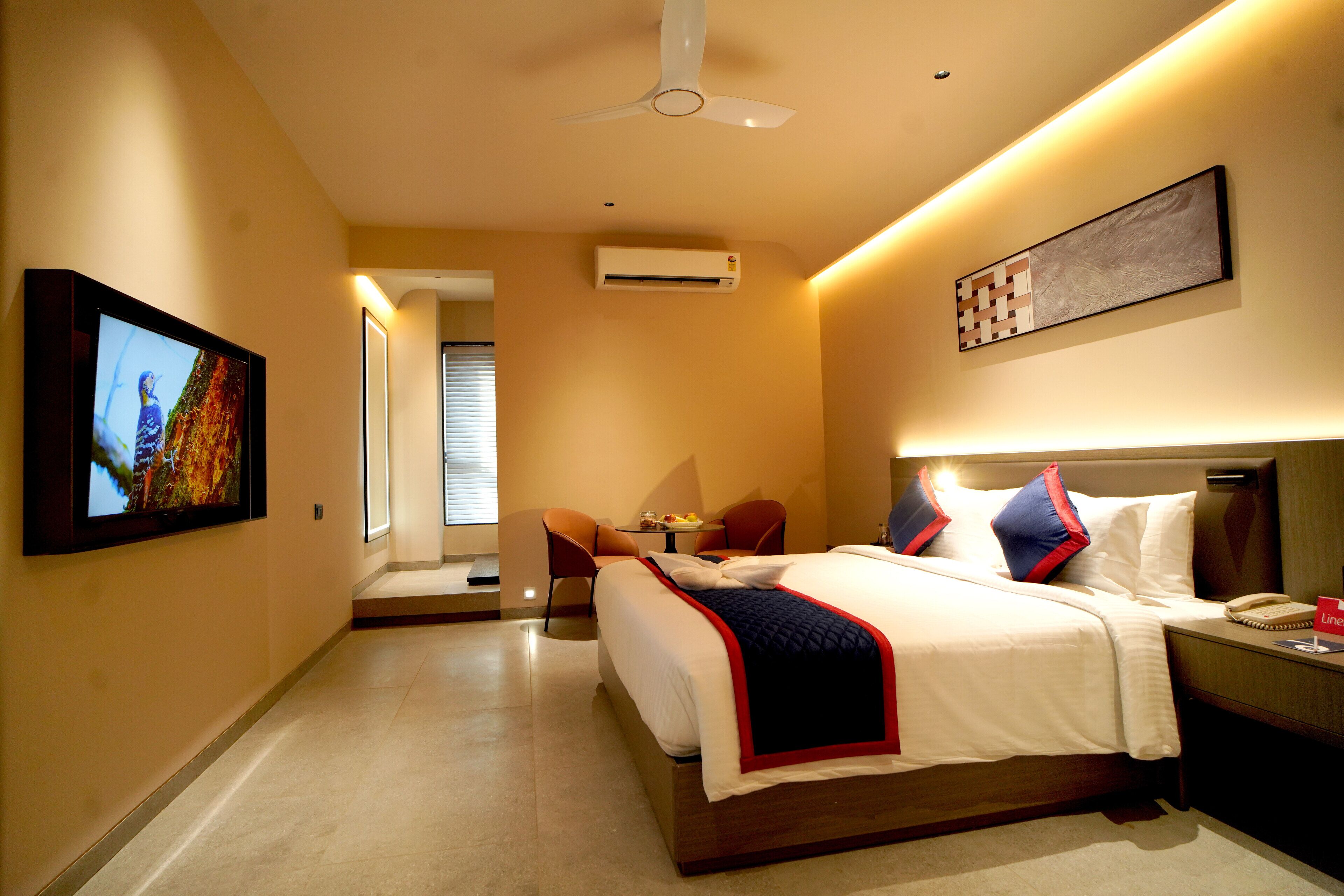 Une chambre d’hôtel comprenant un lit, une table de chevet, une chaise, une télévision, un climatiseur, un ventilateur de plafond et une œuvre d’art au mur.