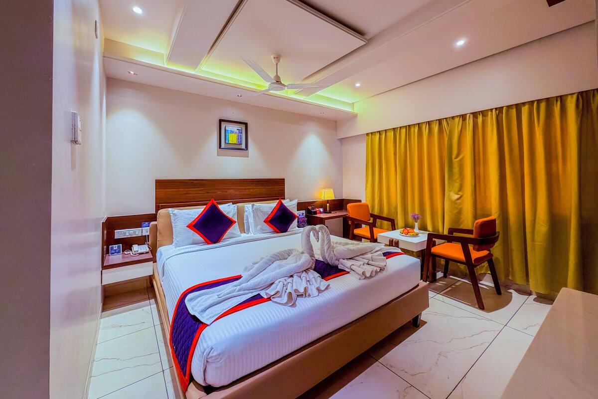 Quarto de hotel com uma cama grande, uma escrivaninha com cadeira, uma pequena mesa com cadeira, um ventilador de teto e uma grande janela com cortinas.