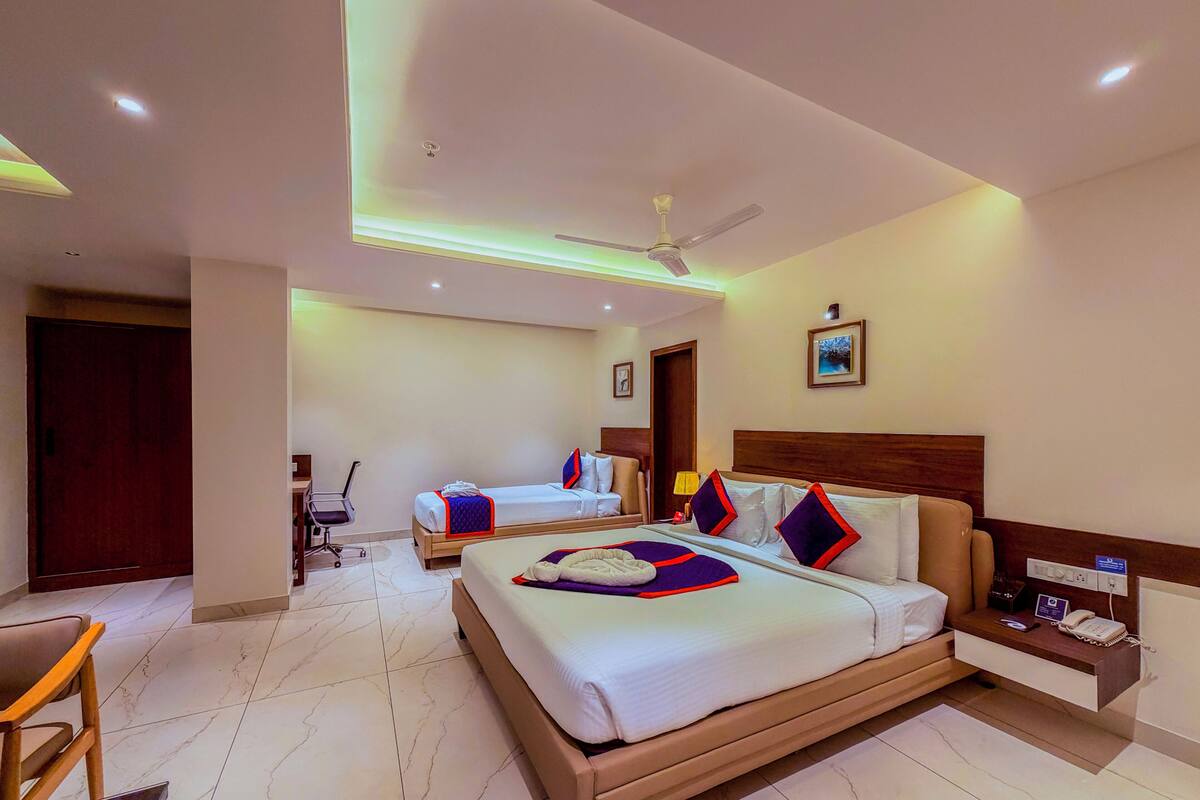 Quarto de hotel com uma cama grande, uma escrivaninha, uma cadeira, um ventilador de teto e uma mesa de cabeceira com telefone e luminária.