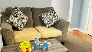 Ein Wohnzimmer mit grauem Sofa, gemusterten Kissen, einem Couchtisch mit Früchten und Getränken sowie einem gerahmten Bild an der Wand.