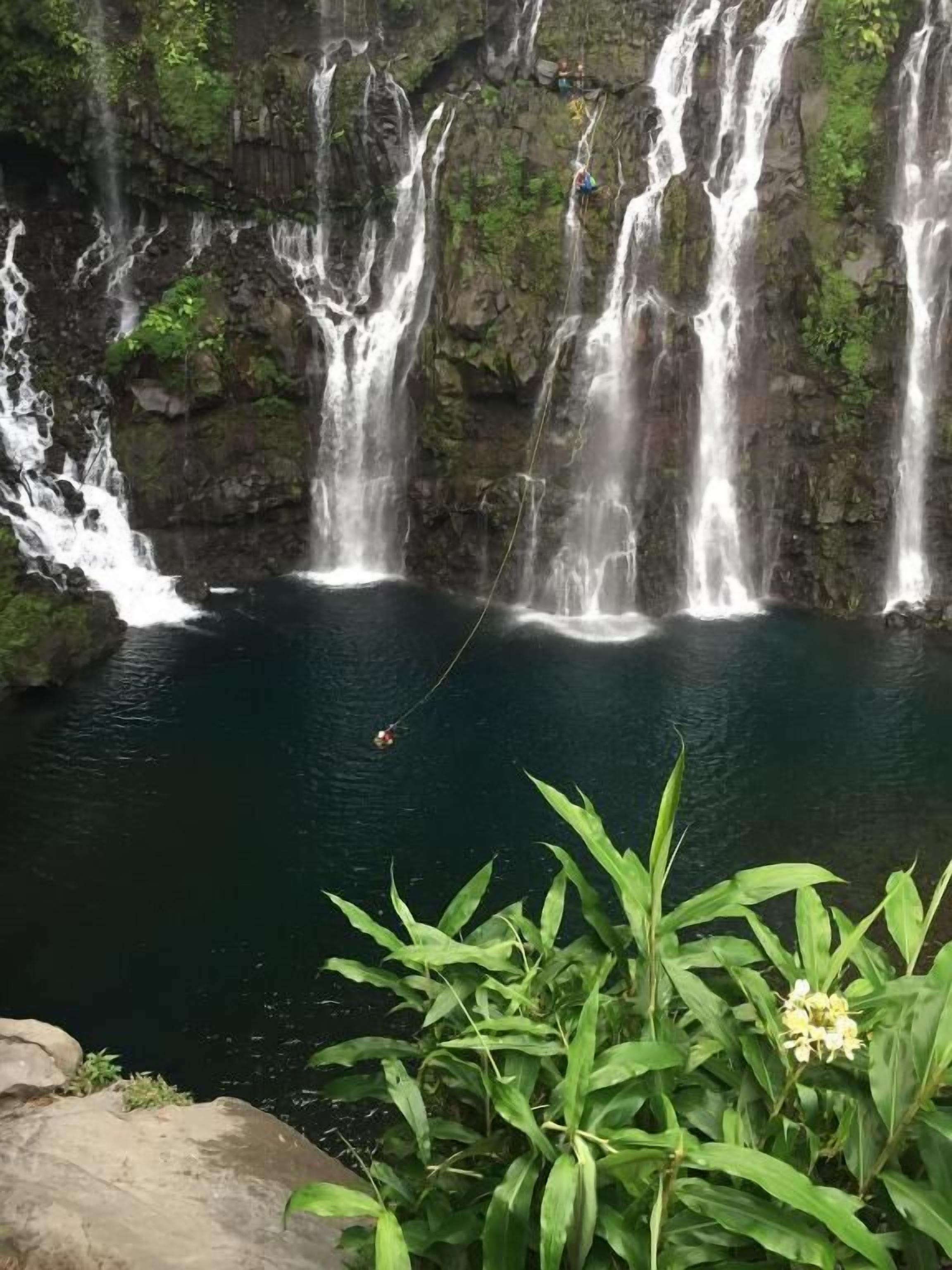 Una cascada con múltiples brazos de agua cayendo en una poza oscura rodeada de acantilados rocosos y vegetación.