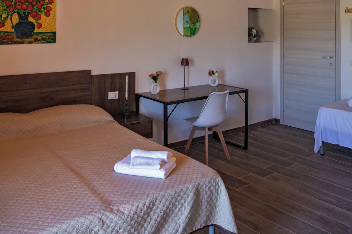 A bedroom with a large bed, a desk, a chair, a small table, a painting, a clock, and a door.