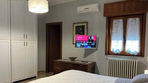Quarto com cama, cômoda, TV na parede e janela com cortinas de renda.