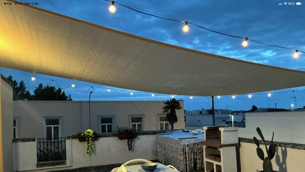 Um pátio externo com mesa e cadeiras, luzes de cordão e plantas em vasos.