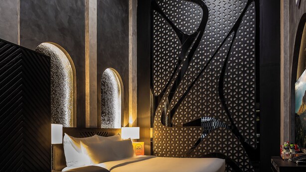 Quarto de hotel moderno com cama, escrivaninha e cadeira. Possui janelas em arco e um painel decorativo na parede.