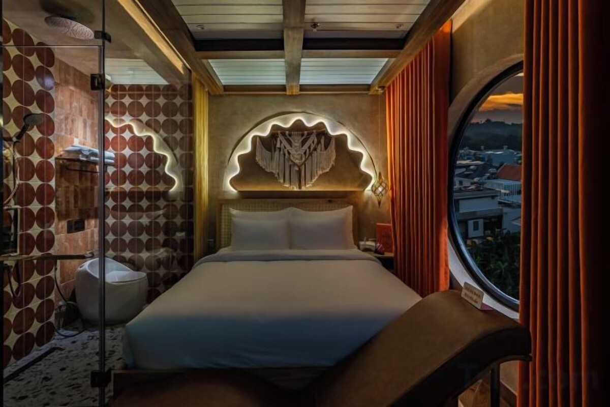 A bedroom with a bed, a circular window, and a city view.