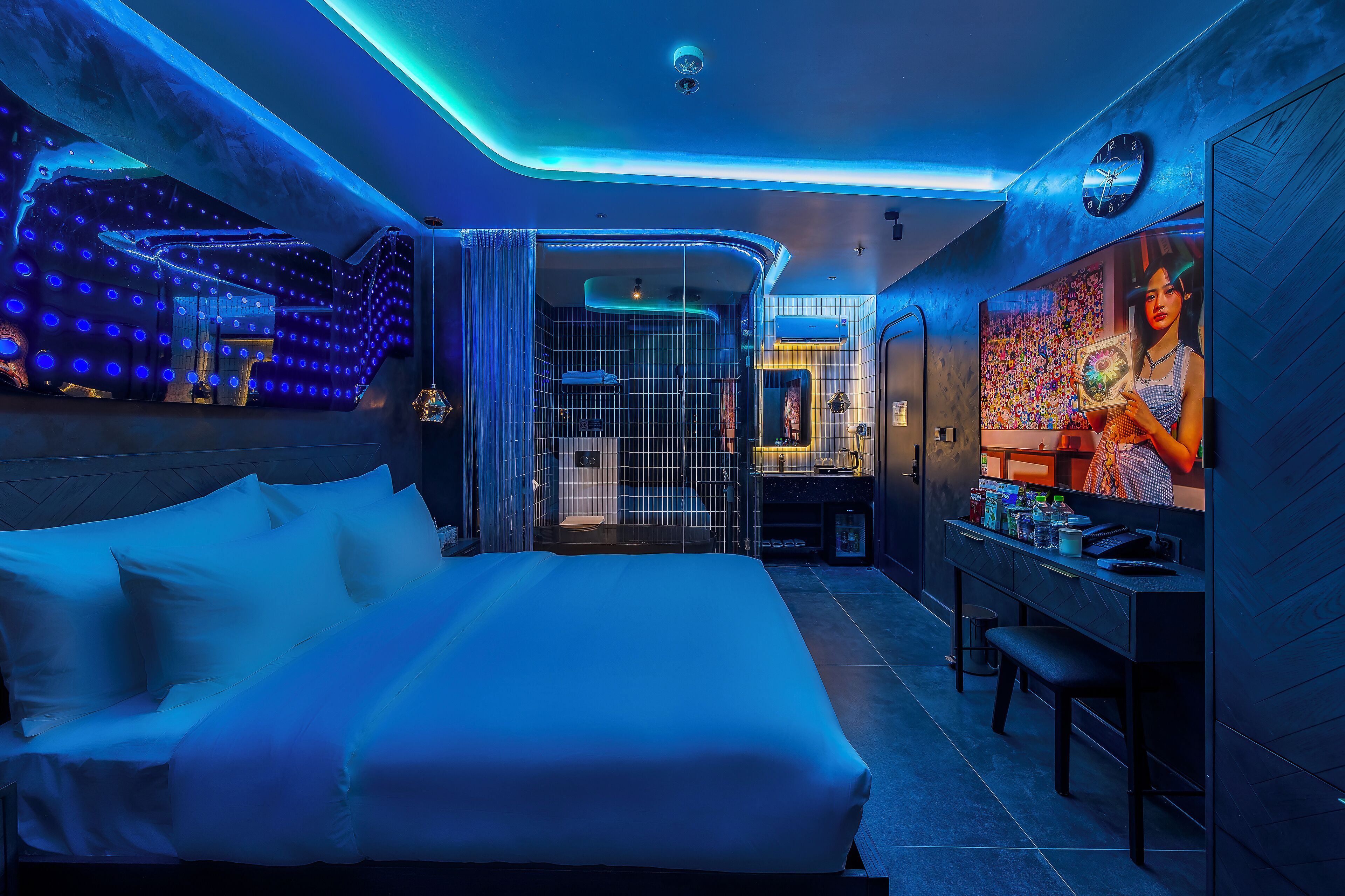 A modern hotel room with a large bed, a desk with a computer, and a bathroom visible through a glass partition.