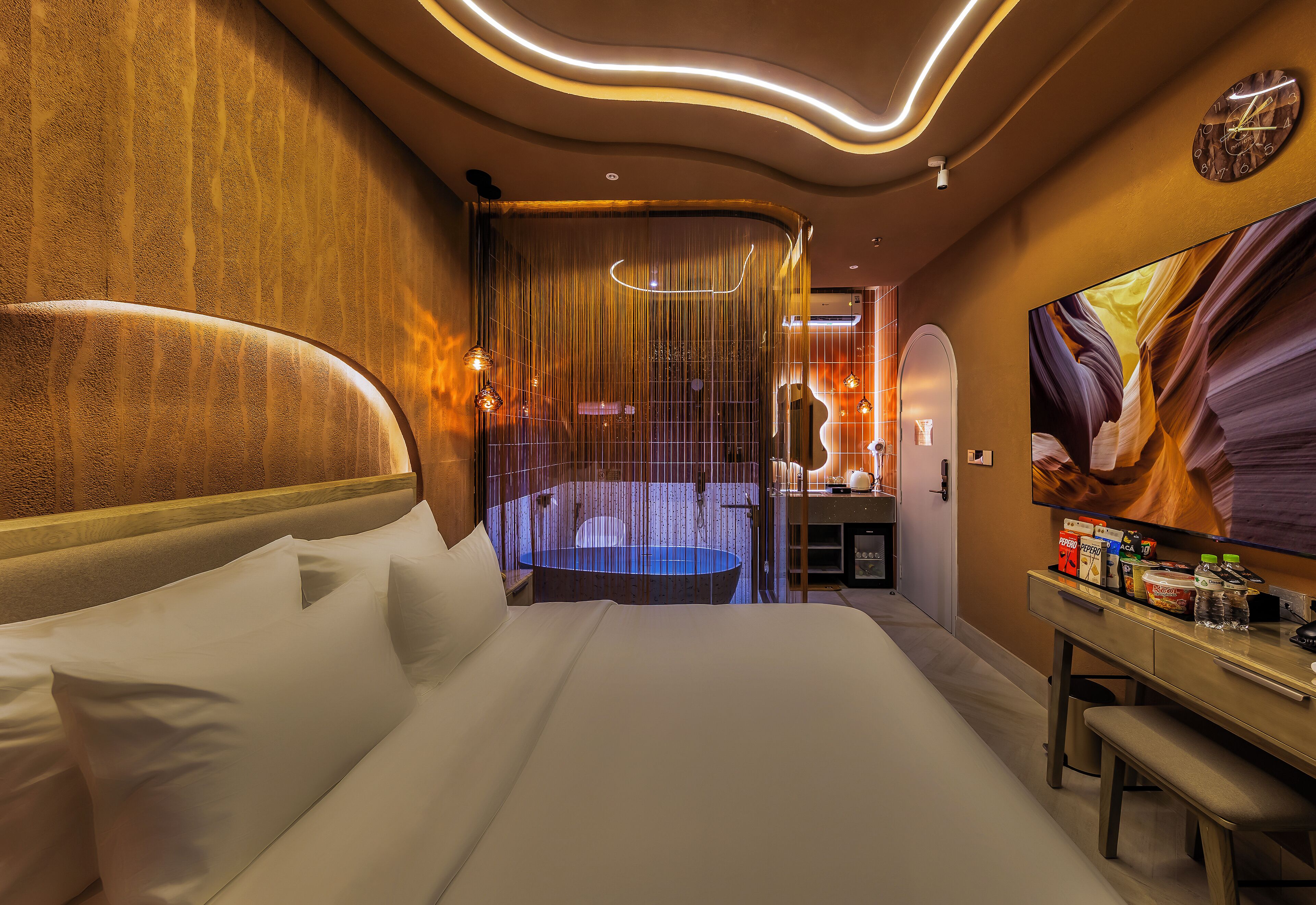 A modern hotel room with a large bed, a desk with a computer, a mirror, and a bathroom with a bathtub.
