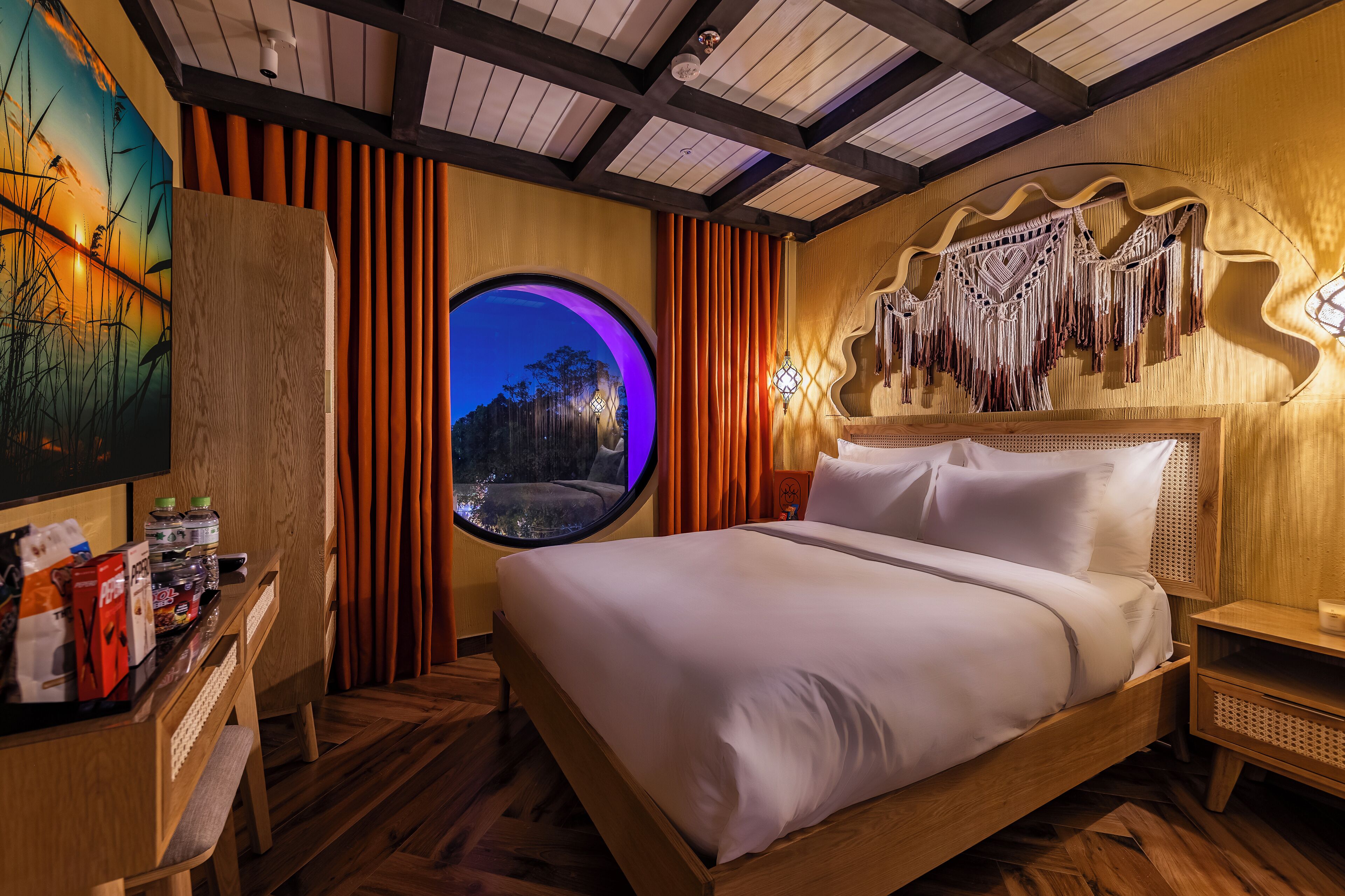 A bedroom with a bed, wooden headboard, a round window, and a wall-mounted macrame piece.