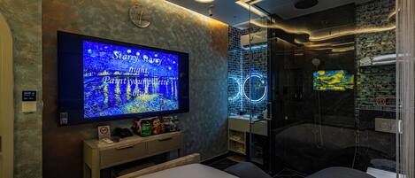 Una habitación de hotel moderna con un televisor de pantalla plana grande, cabina de ducha de vidrio y cama con cabecero.