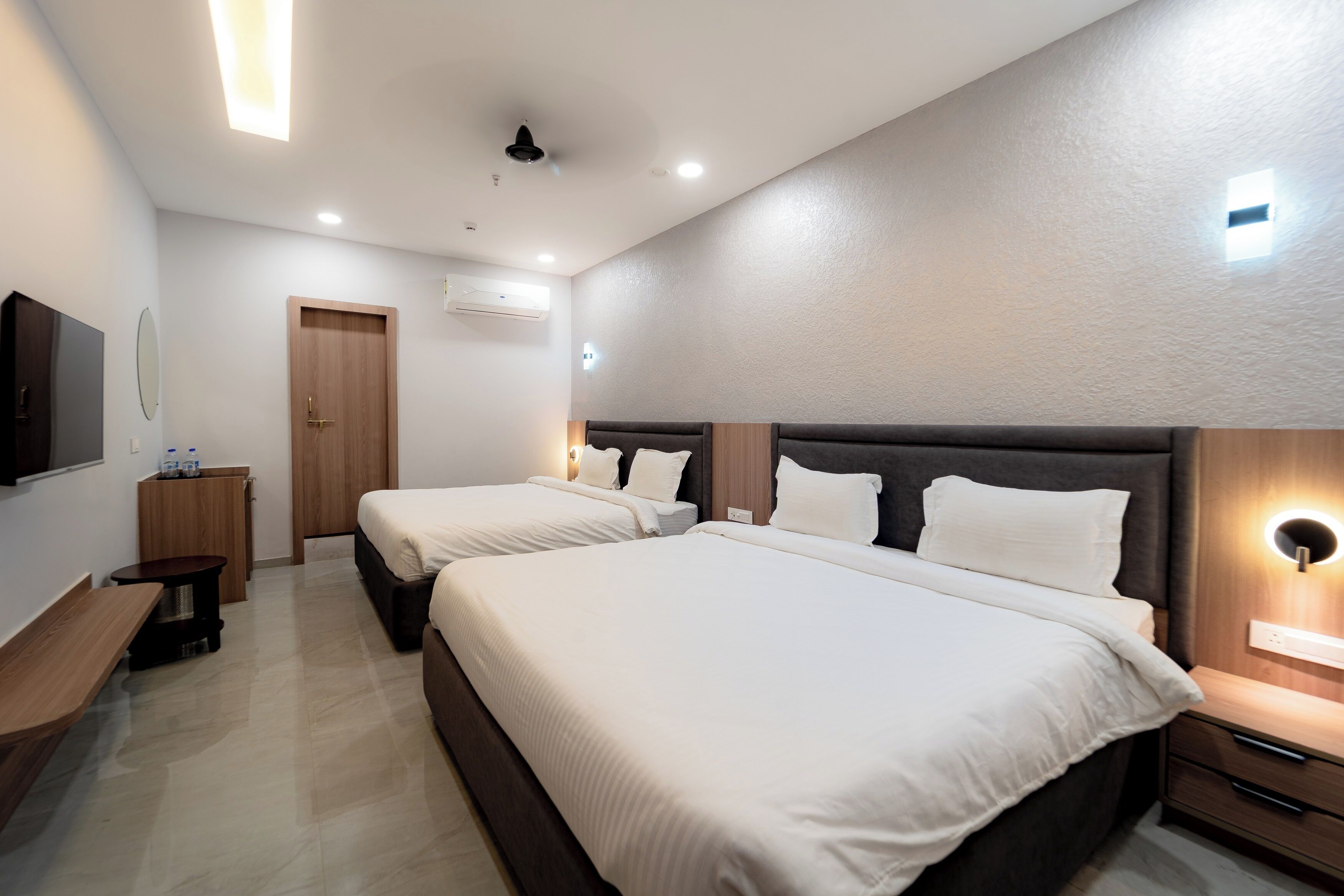 A modern hotel room with two beds, a wooden wardrobe, a small table, and a wall-mounted TV.