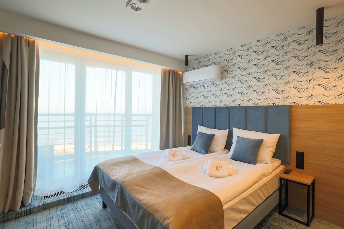 Hotellihuone, jossa on suuri sänky, näkymä ulos, ilmastointilaite ja pieni yöpöytä.