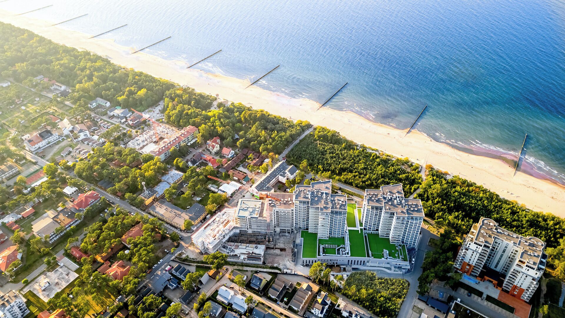 An aerial view of a coastal residential area with a large building complex featuring multiple high-rise apartments, a central green space, and a beach in the background.