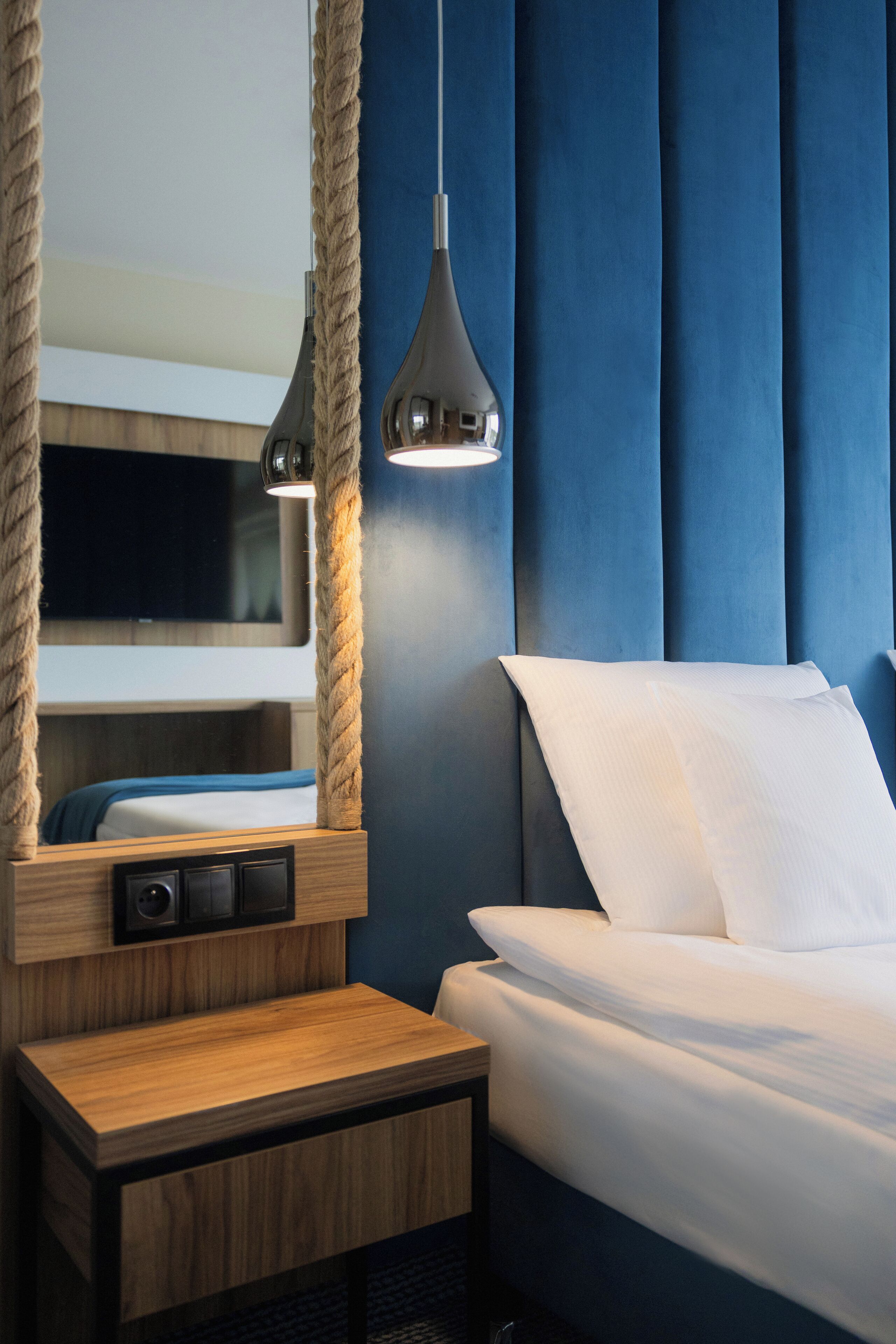 A hotel room with a bed, a wooden nightstand, a mirror, and a rope-wrapped light fixture.