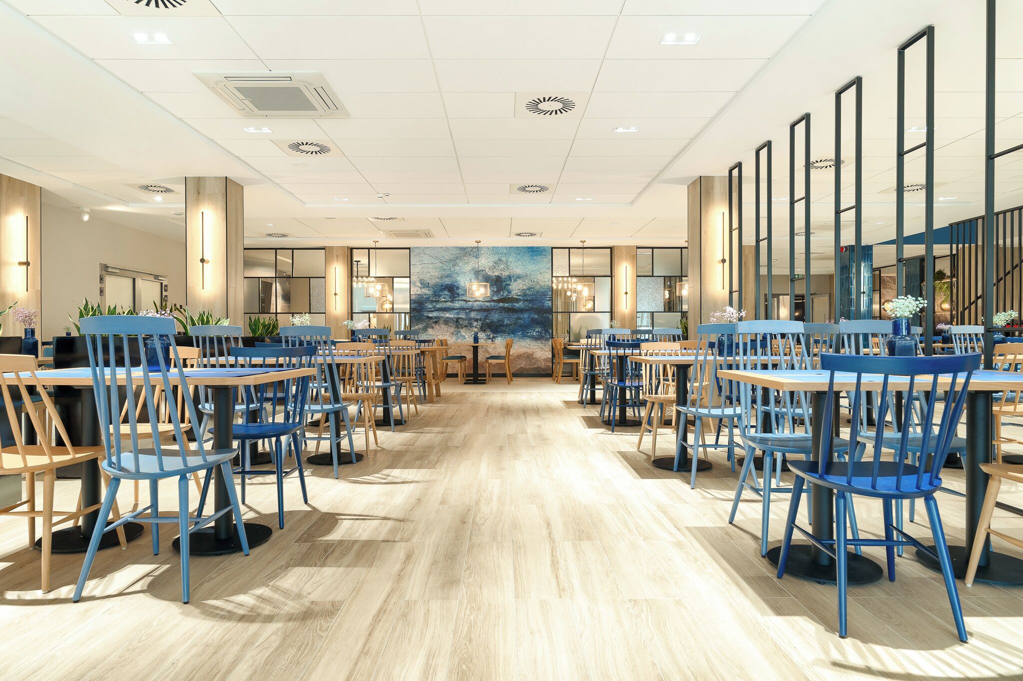 A spacious dining area with multiple tables and chairs, a large mural on the back wall, and modern lighting fixtures.