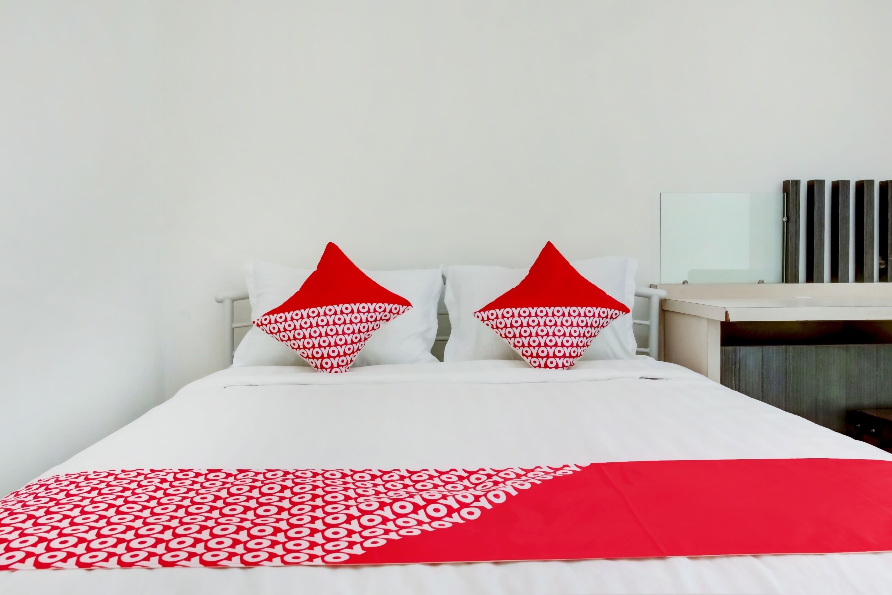 A bed with white bedding and red pillows, a red throw blanket, and a wooden nightstand with a mirror.