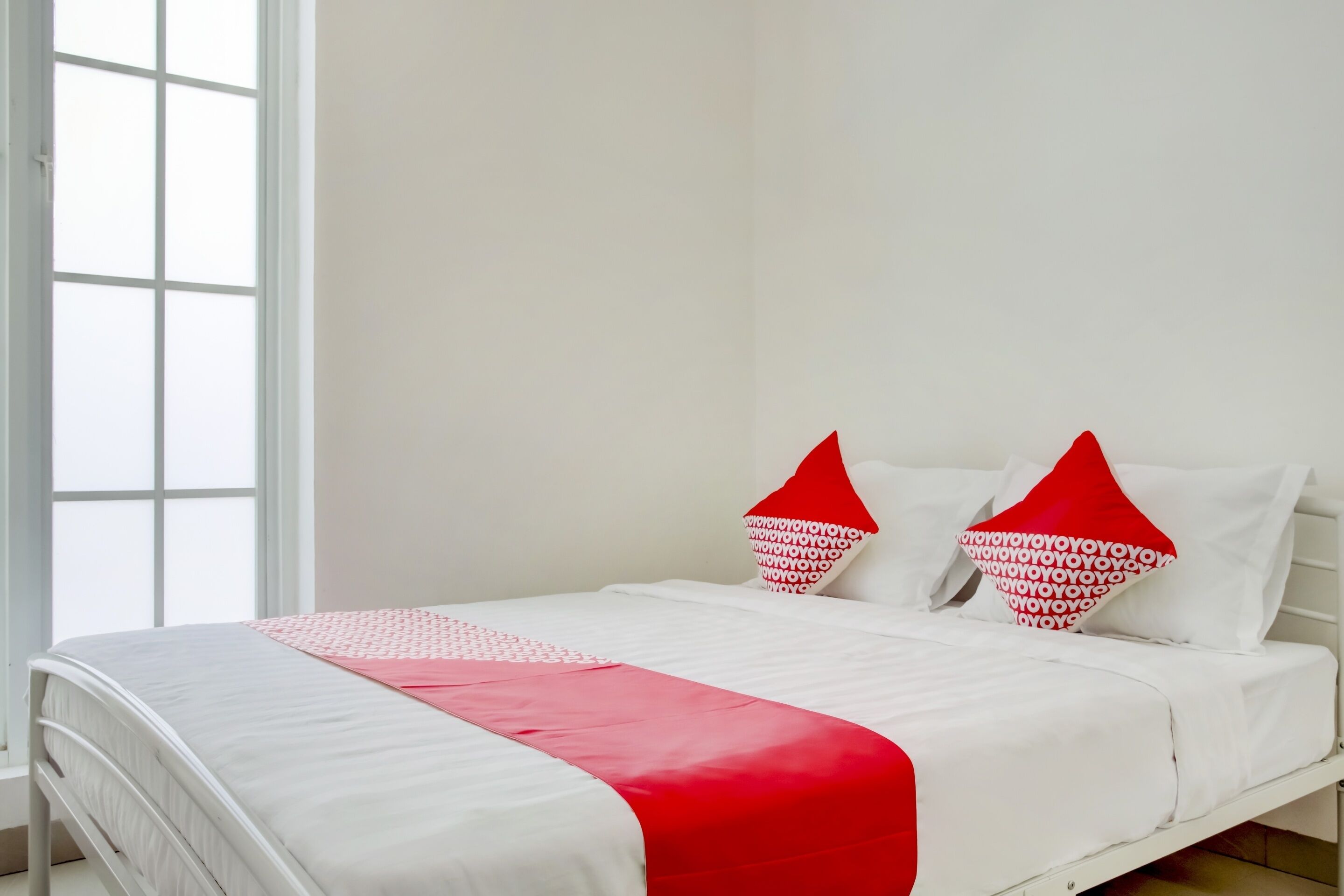 A neatly made bed with white and red bedding, a window with white frames, and a plain wall.