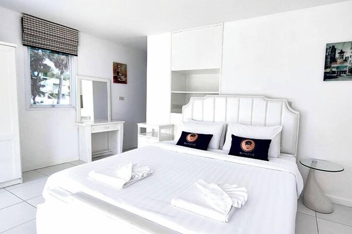 Une chambre d’hôtel moderne avec un grand lit, des meubles blancs, une petite table de chevet et une fenêtre avec des stores.