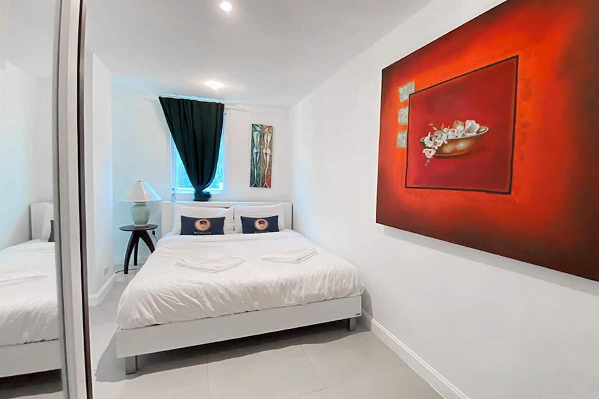 Une chambre moderne avec un lit blanc, une table de chevet avec une lampe, une fenêtre avec un rideau et un tableau abstrait rouge accroché au mur.