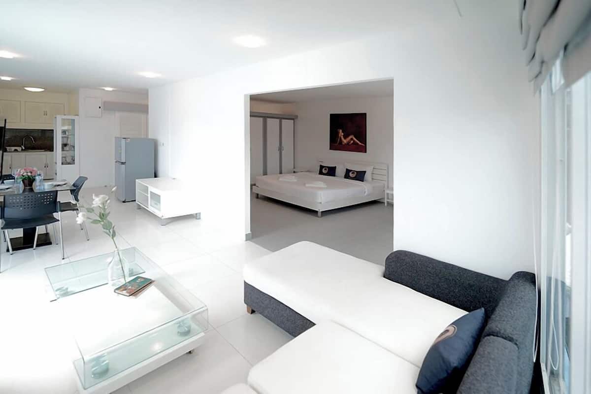 Un salon moderne avec une table basse en verre, un canapé gris et un lit blanc en arrière-plan.