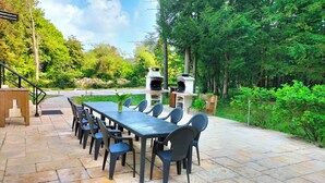 Une terrasse extérieure avec une longue table et des chaises, entourée de verdure et agrémentée de deux fours à pizza en pierre.