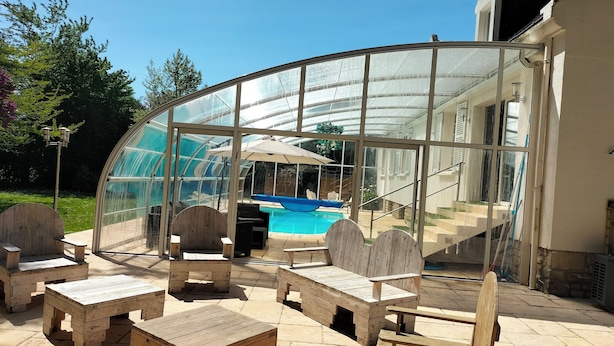 Une terrasse fermée par des baies vitrées, avec une piscine, des meubles en bois et un escalier menant à un bâtiment.