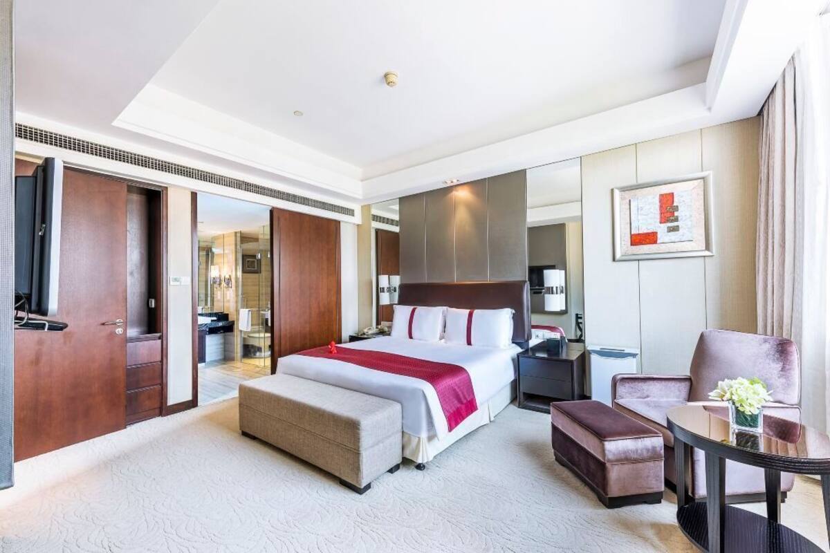 A hotel room with a large bed, a sitting area with a chair and table, a wardrobe, and a mirror.