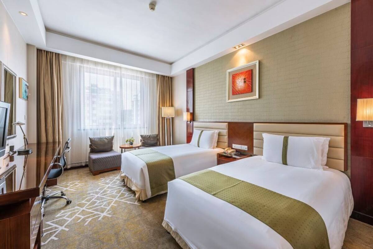 A hotel room with two beds, a desk, a chair, a TV, a lamp, and a window with curtains.
