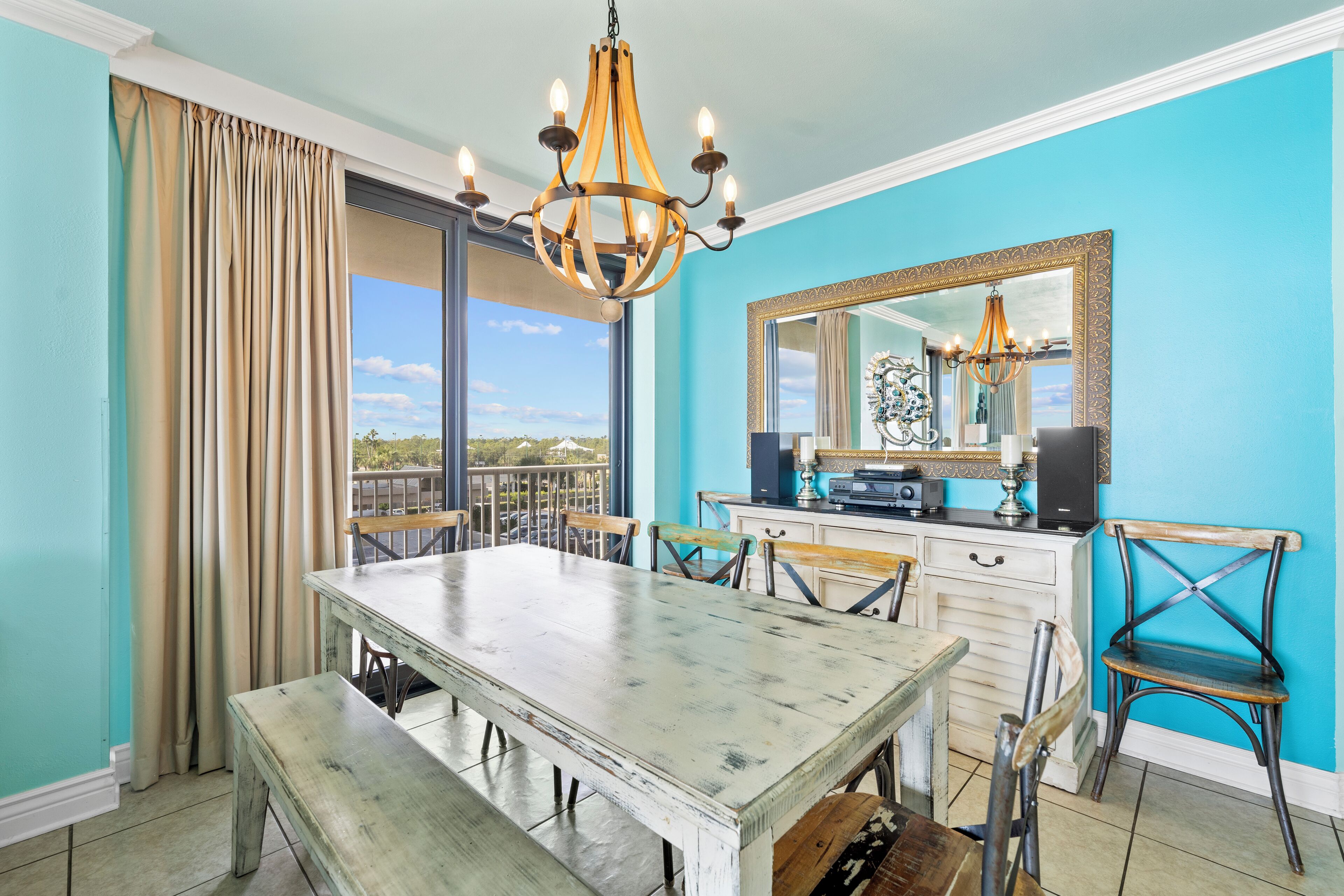 A dining area with a large table, chairs, a chandelier, a mirror, and a view through large windows.