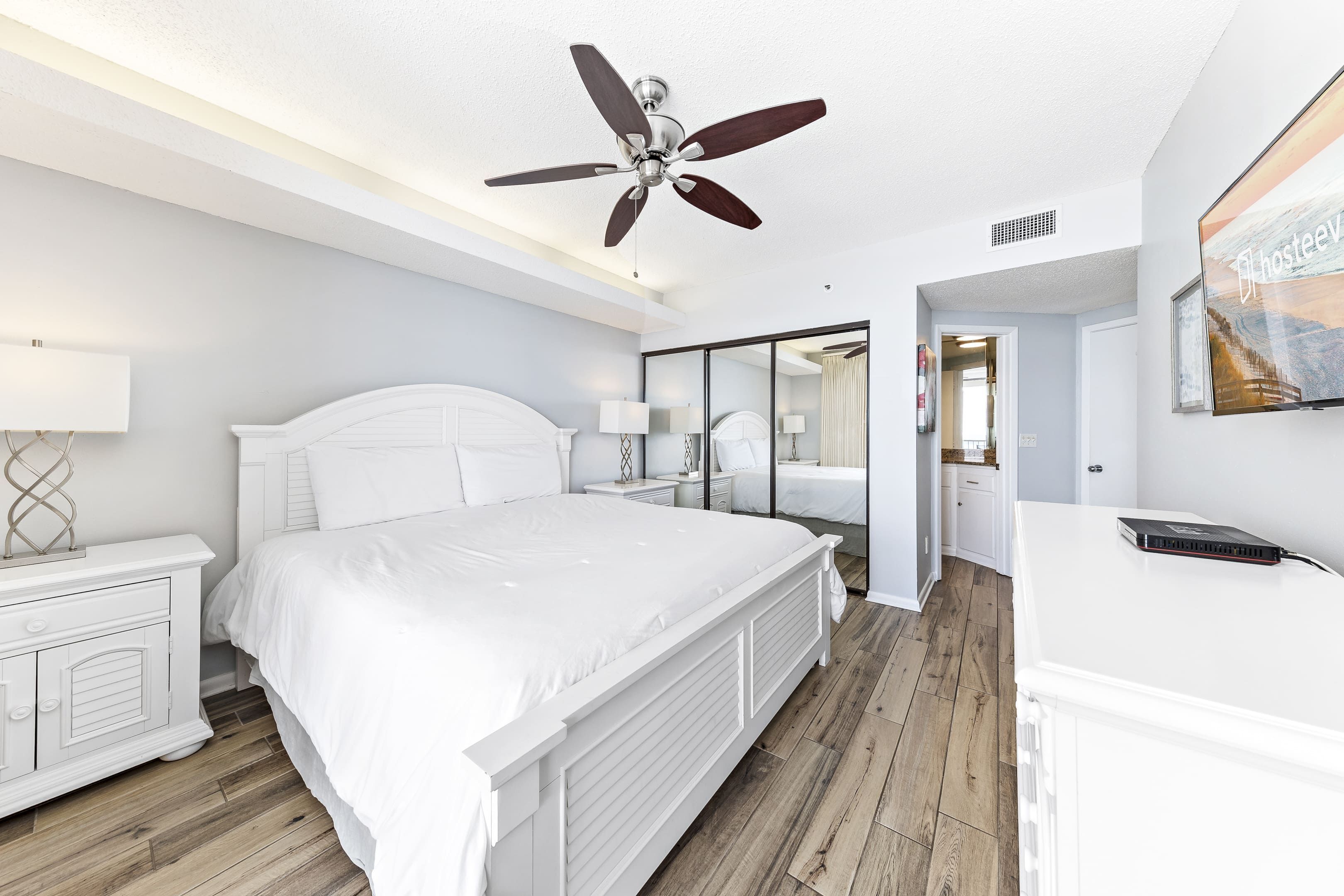 Un dormitorio moderno con una cama grande, un ventilador de techo, una mesita de noche blanca y un espejo.
