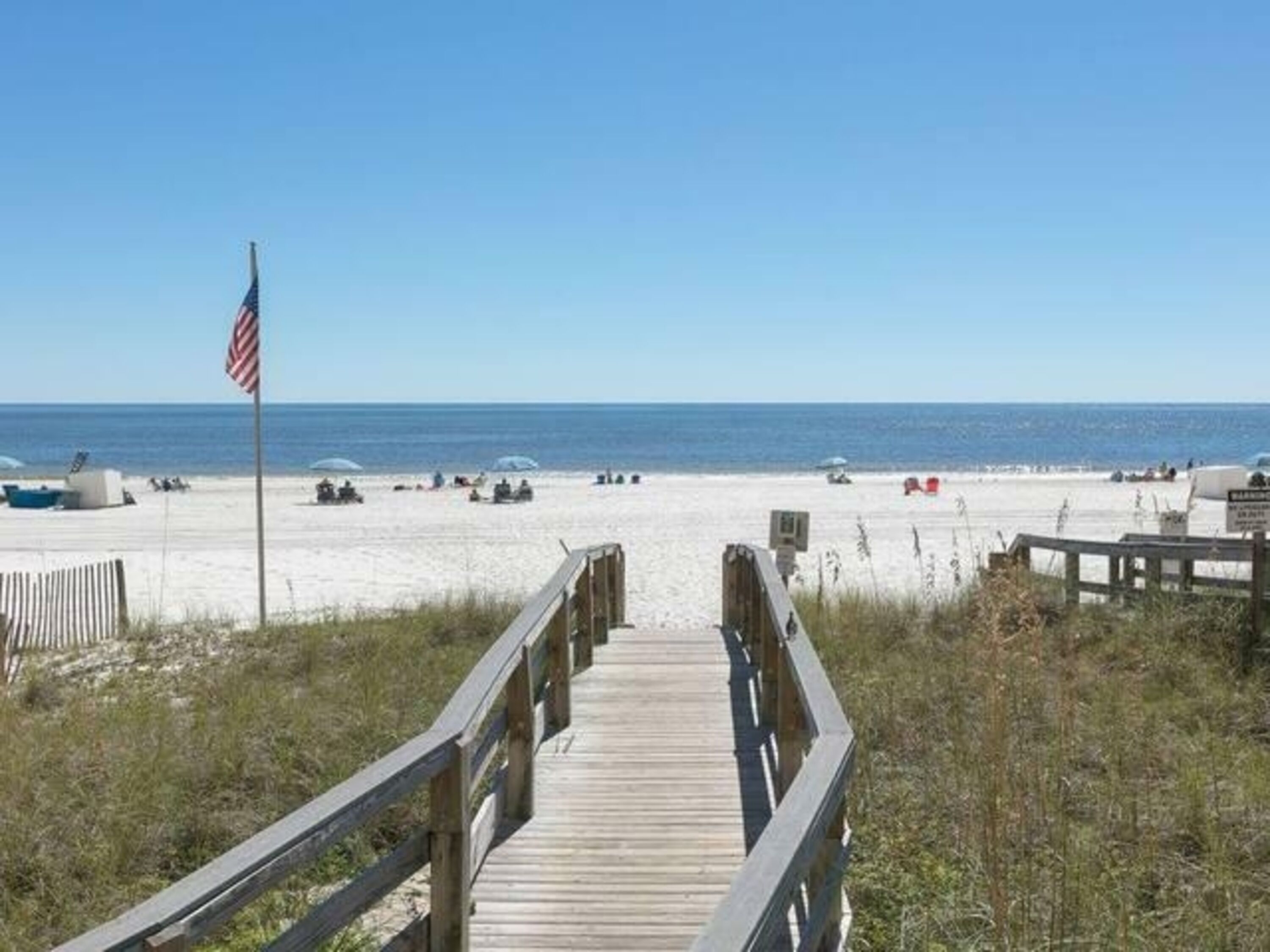 A wooden boardwalk leading to a sandy beach with an American flag, clear skies, and the ocean in the background.
