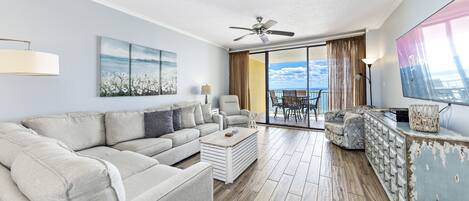 Una sala de estar moderna con un sofá seccional color beige, una mesa de centro y un aparador decorativo. Hay un ventanal con vista al océano y un ventilador de techo.