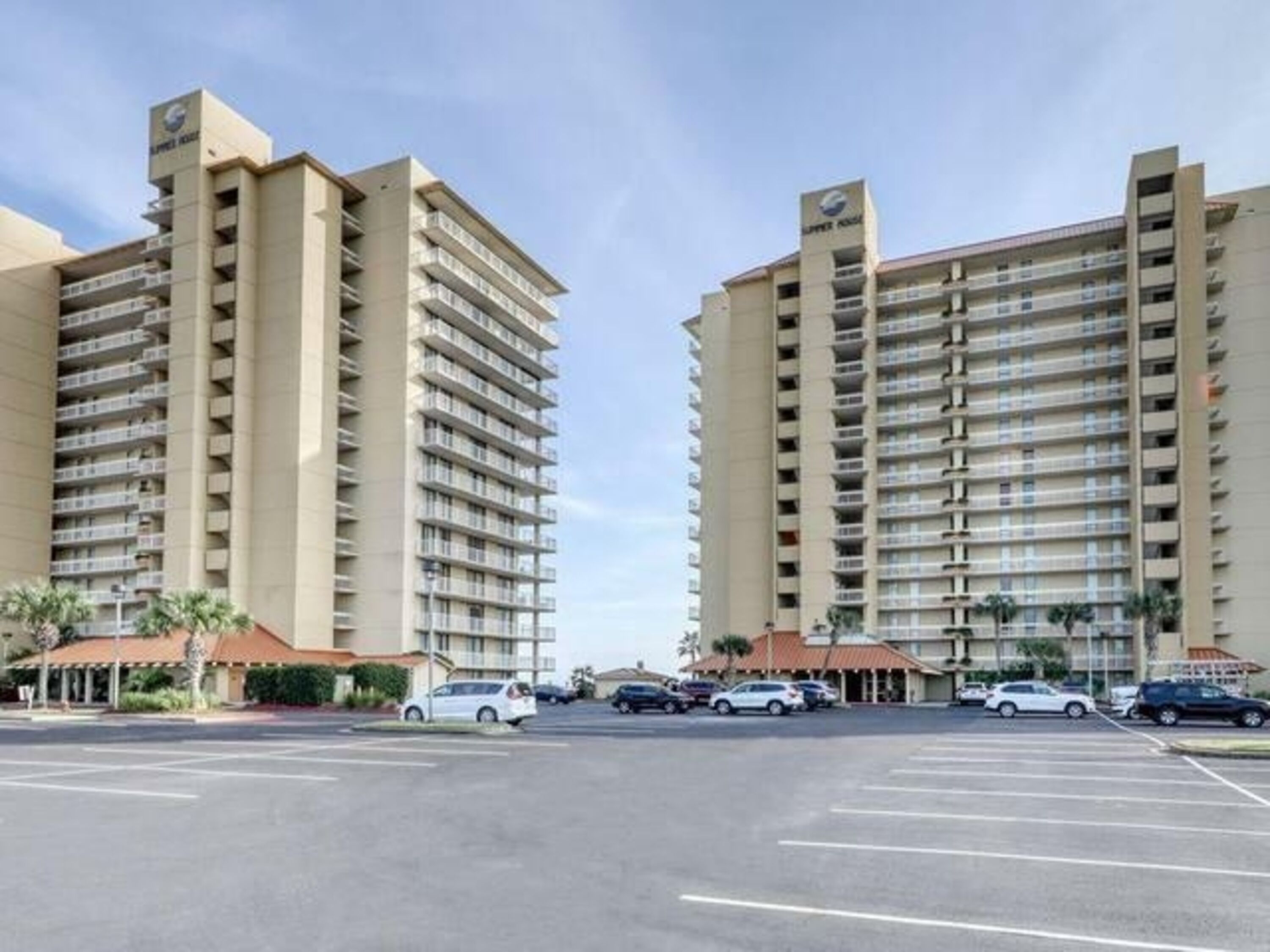 A large parking lot with multiple cars, two tall buildings with balconies, and a clear sky.
