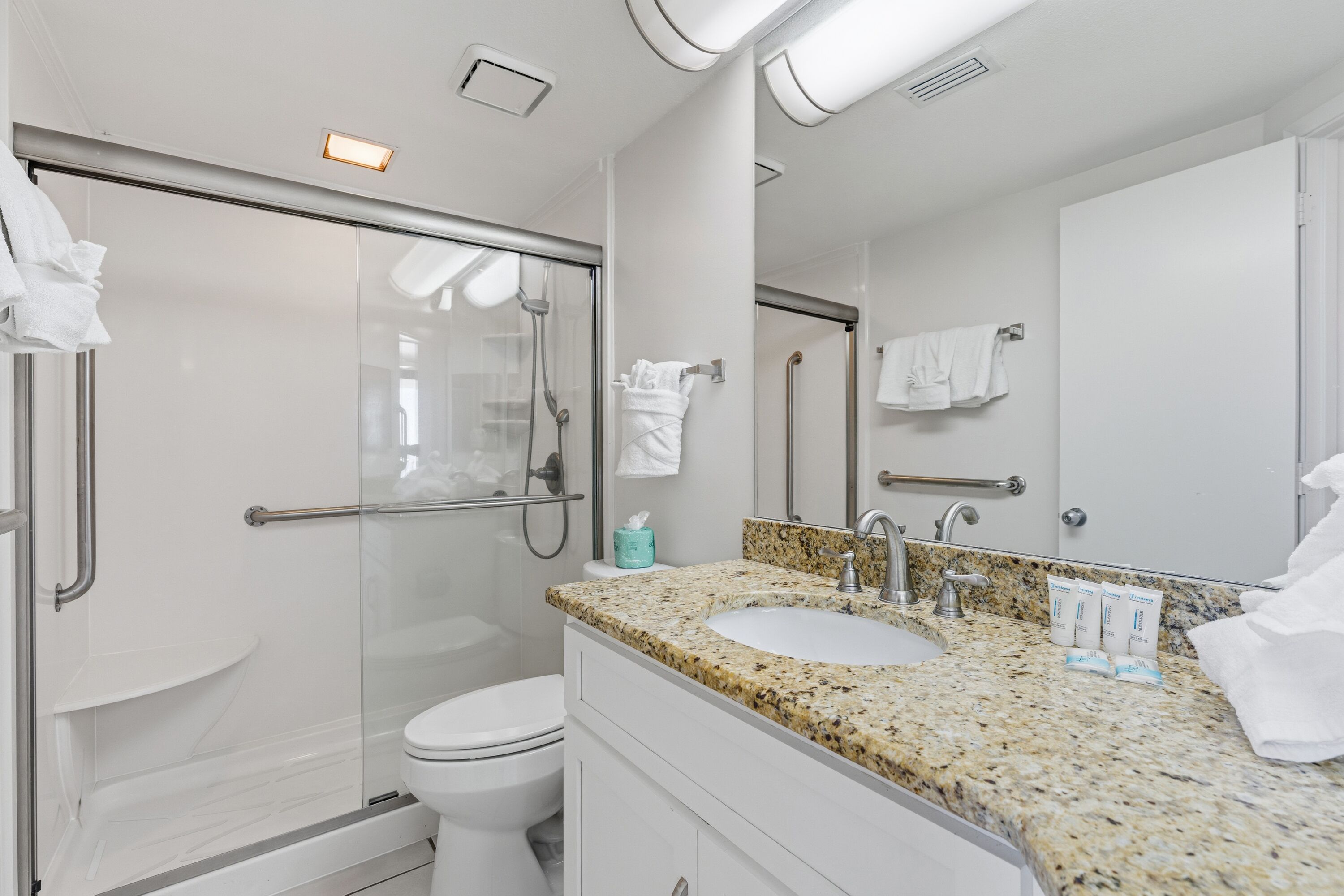 Un baño moderno con cabina de ducha de vidrio, inodoro blanco, lavabo con encimera de granito y un espejo grande.