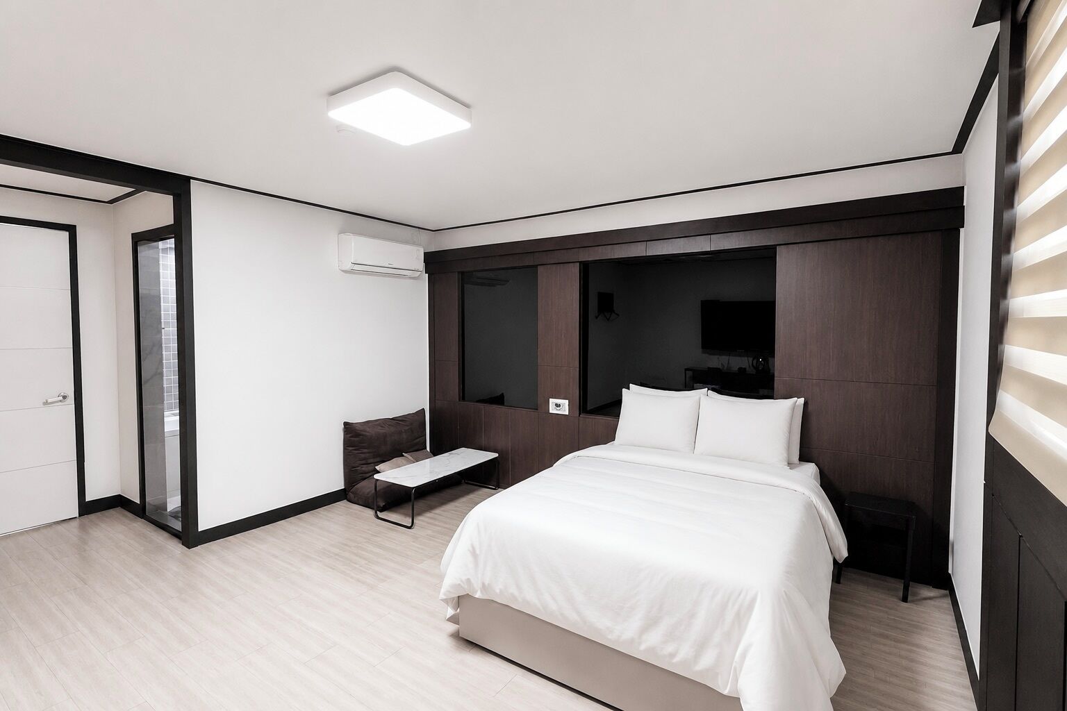 A modern hotel room with a large bed, a small bedside table, a built-in TV, and a large window.