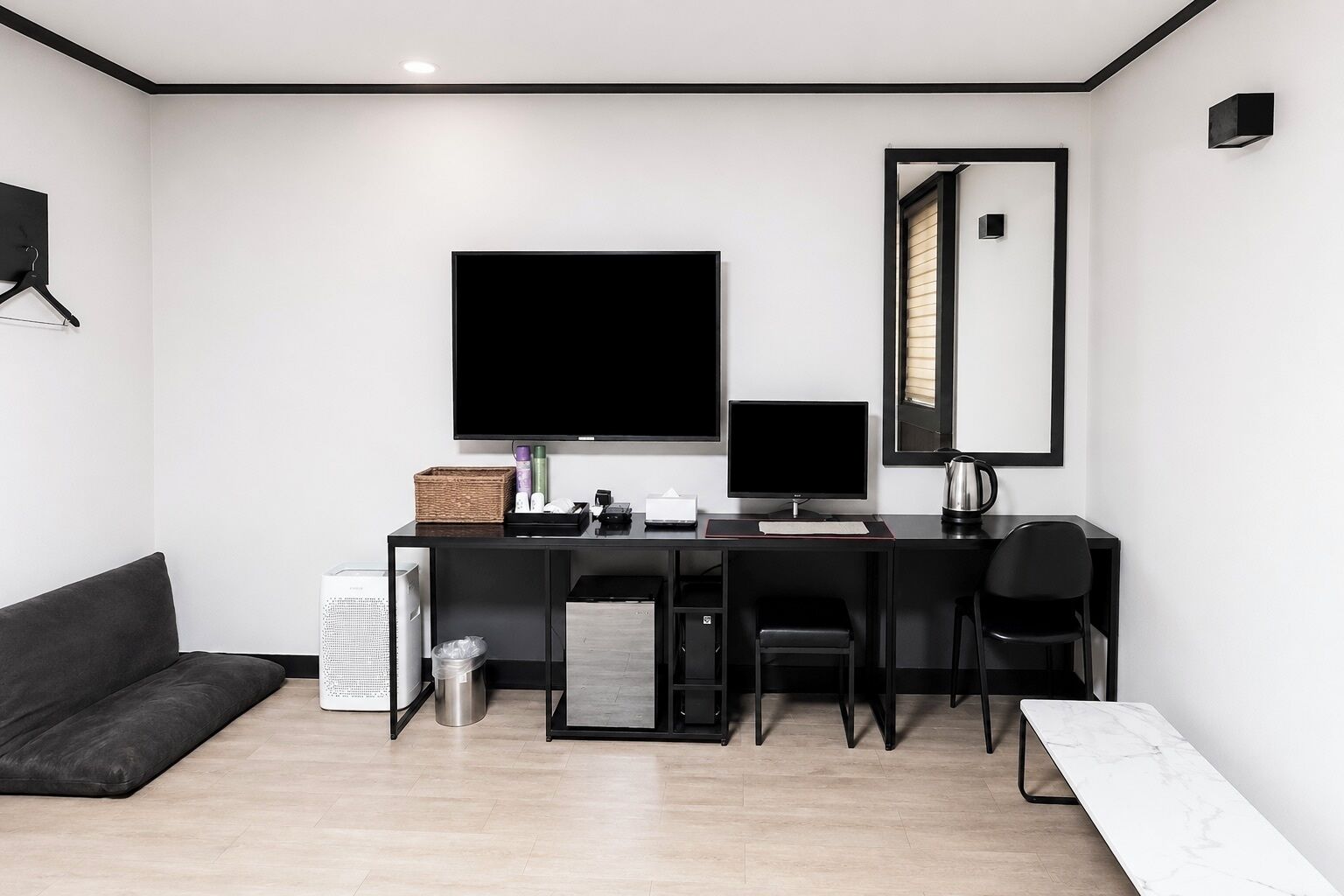 A modern hotel room with a black sofa, a desk with a computer and a flat-screen TV, a small trash bin, and a mirror on the wall.