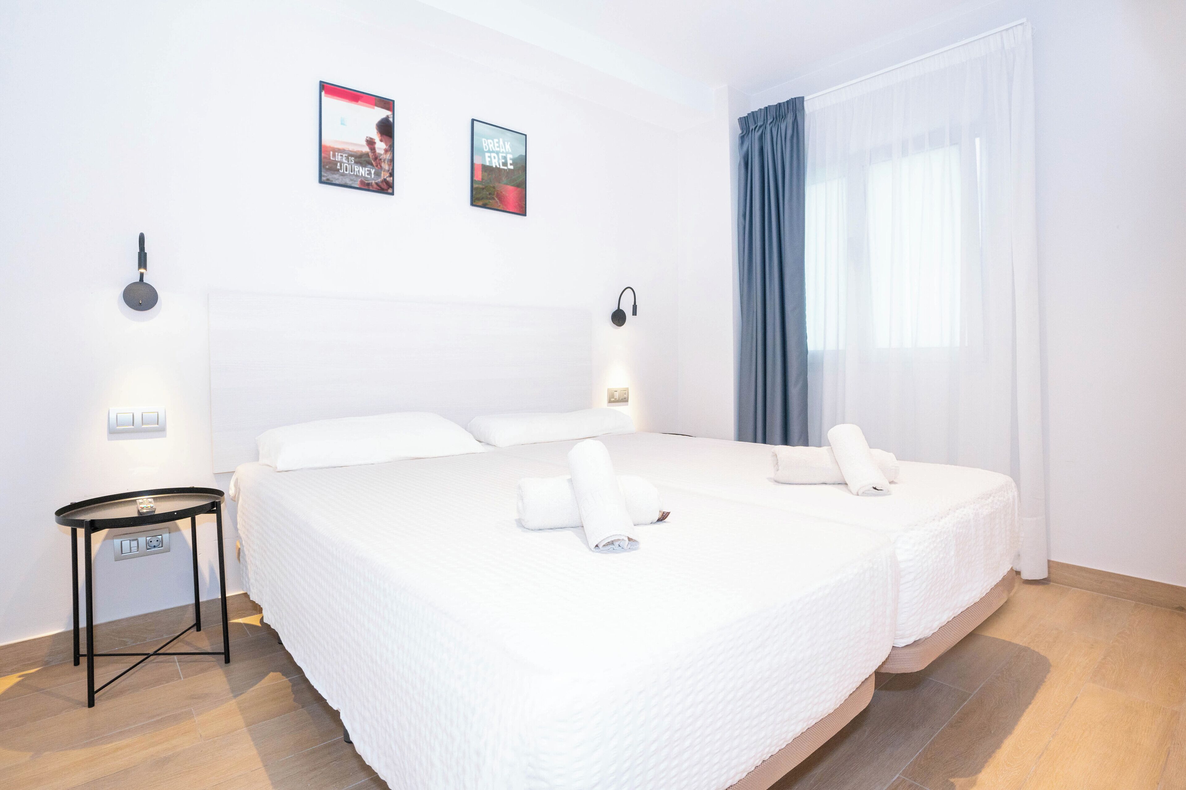 Quarto de hotel com uma cama grande, duas luminárias fixadas na parede, uma mesinha com tomada e duas fotos emolduradas.