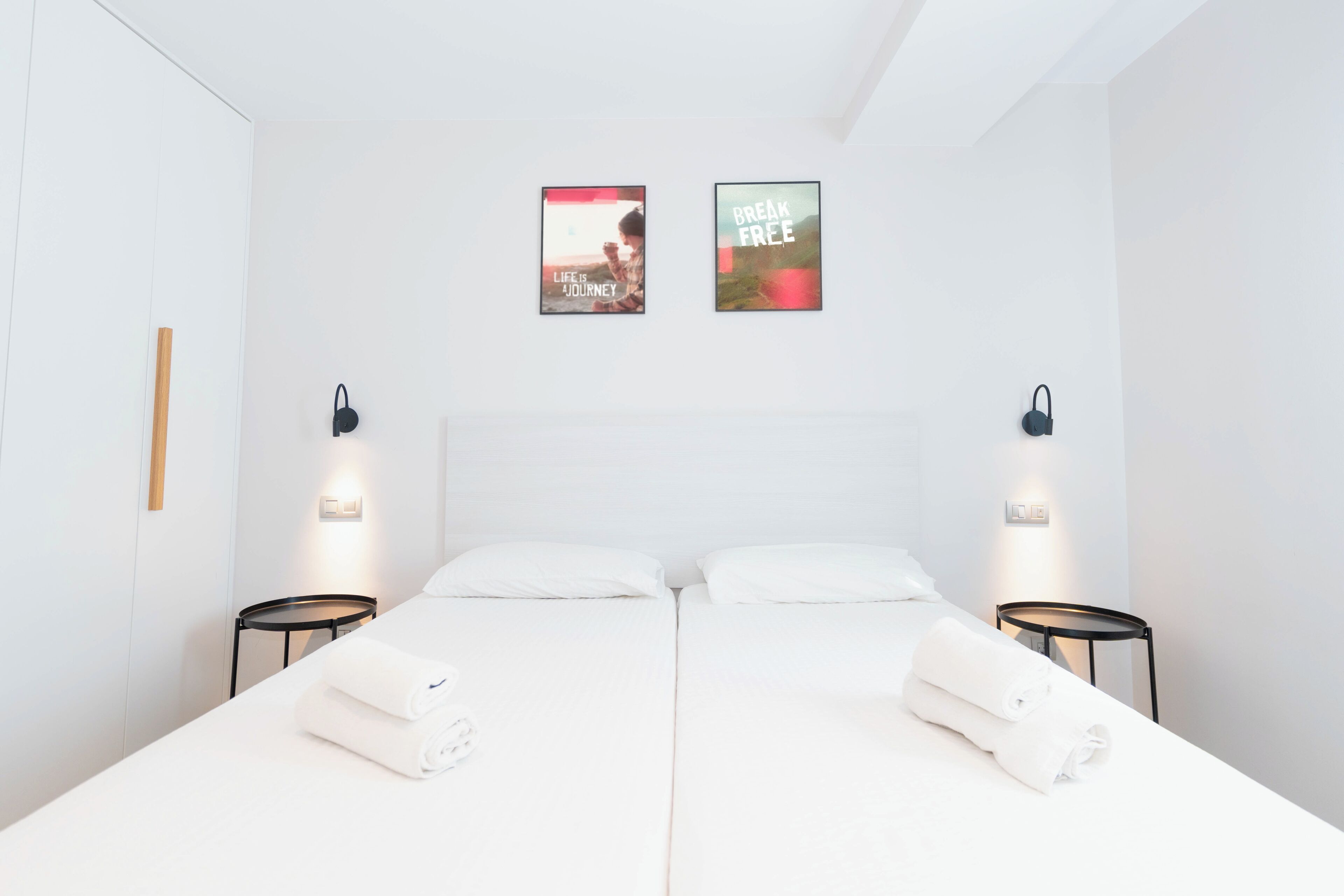 Quarto de hotel com duas camas, roupa de cama e toalhas brancas. Duas mesas de cabeceira fixadas na parede com luminárias. Duas imagens motivacionais estão expostas acima das camas.