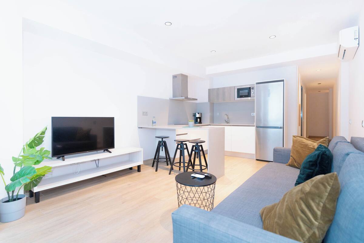 Uma sala de estar moderna com uma televisão de tela plana, um sofá, uma mesa de centro e uma cozinha com geladeira e micro-ondas.