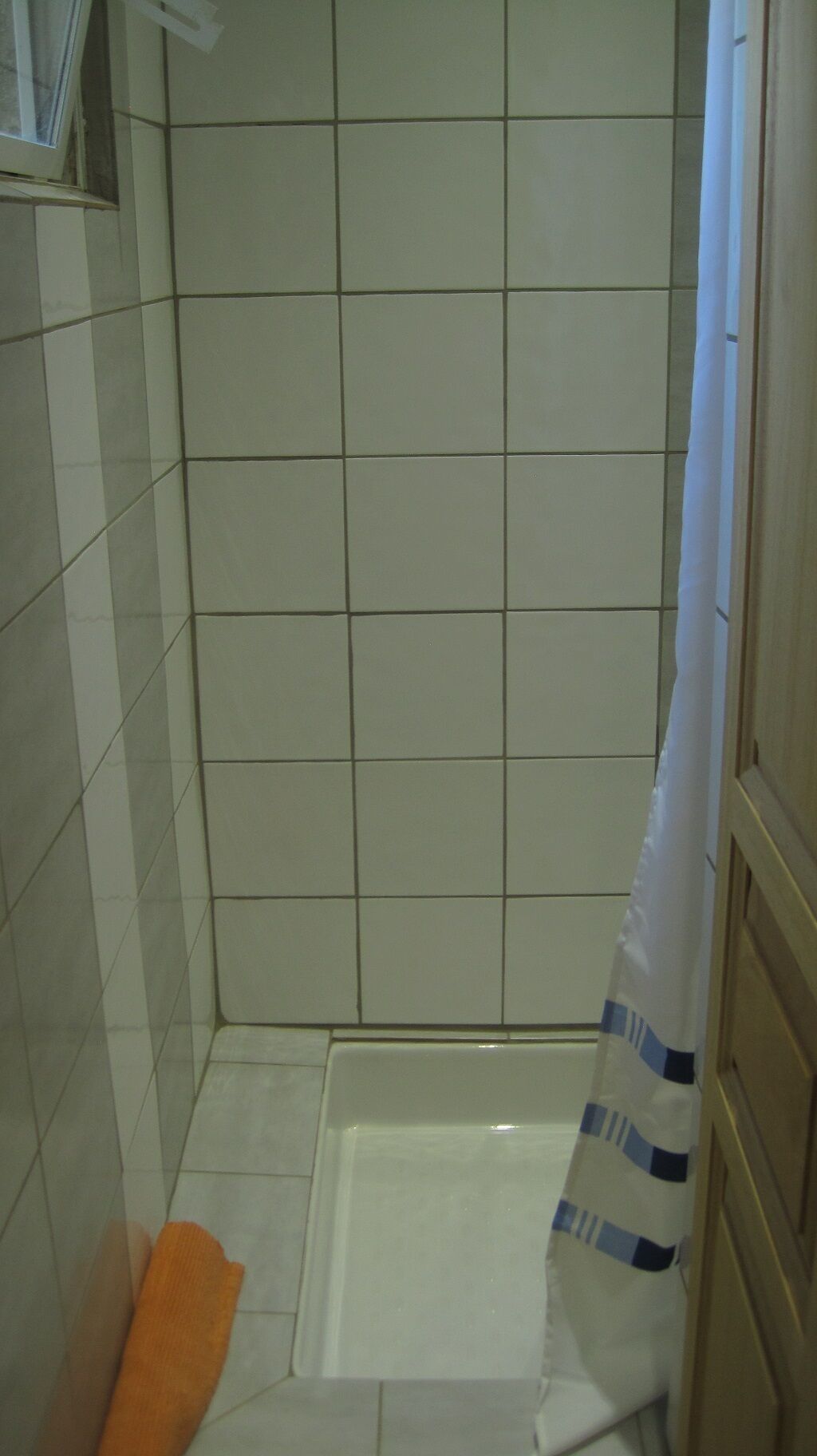 Een kleine, betegelde douchecabine met een witte douchegordijn en een enkele oranje handdoek op de vloer.
