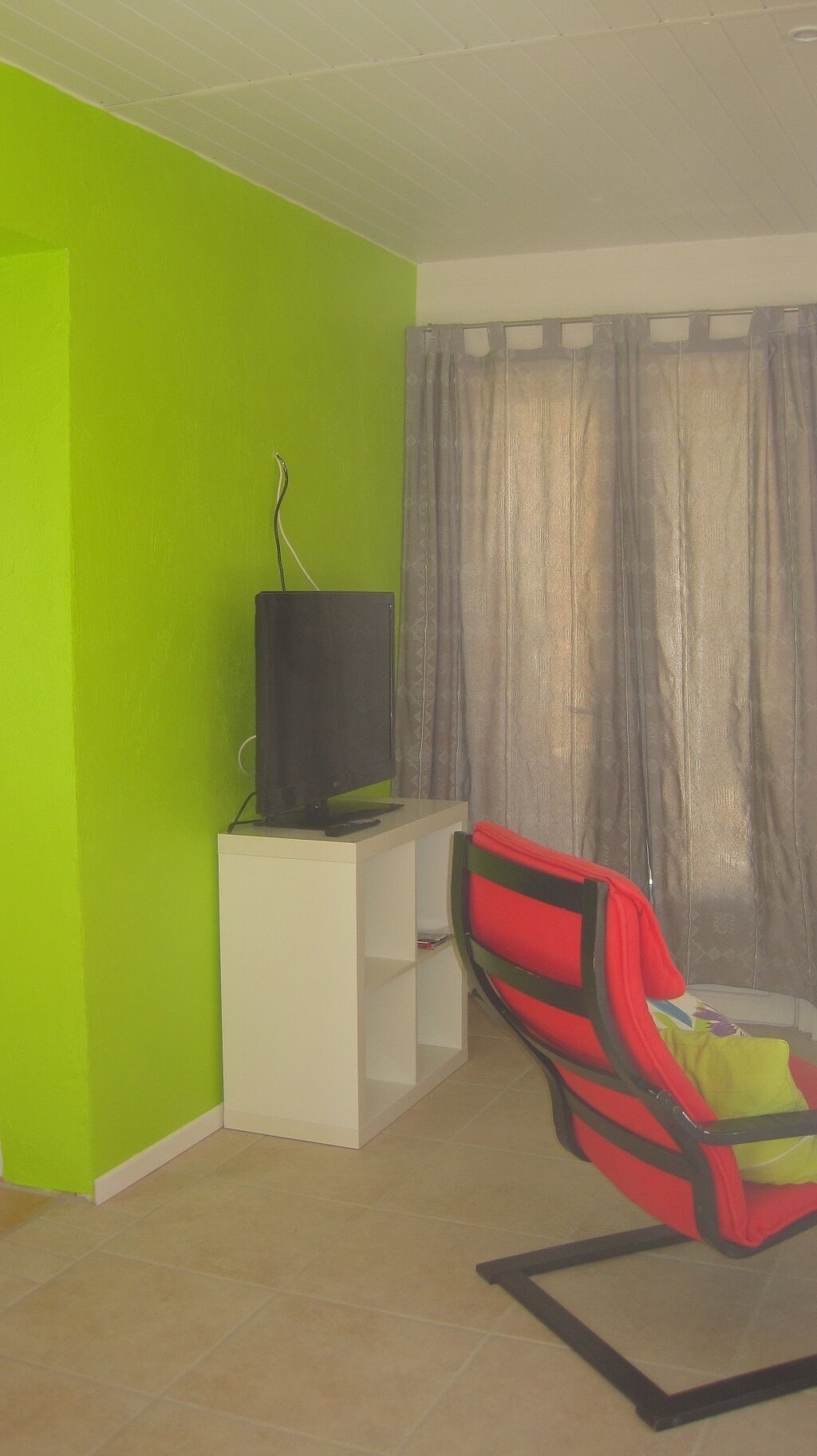 Ein Wohnzimmer mit grüner Wand, einer weißen Unterhaltungseinheit, einem Fernseher, einem rot-schwarzen Sessel und einem grauen Vorhang.