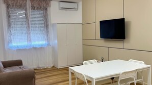 Un salon moderne avec une table et des chaises blanches, un téléviseur à écran plat fixé au mur et un canapé marron.