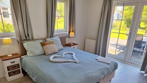 A bedroom with a bed, nightstands, a lamp, a heart-shaped towel on the bed, and a view through the window. - Charming Sauna Home Near Alkmaar (West-Graftdijk)