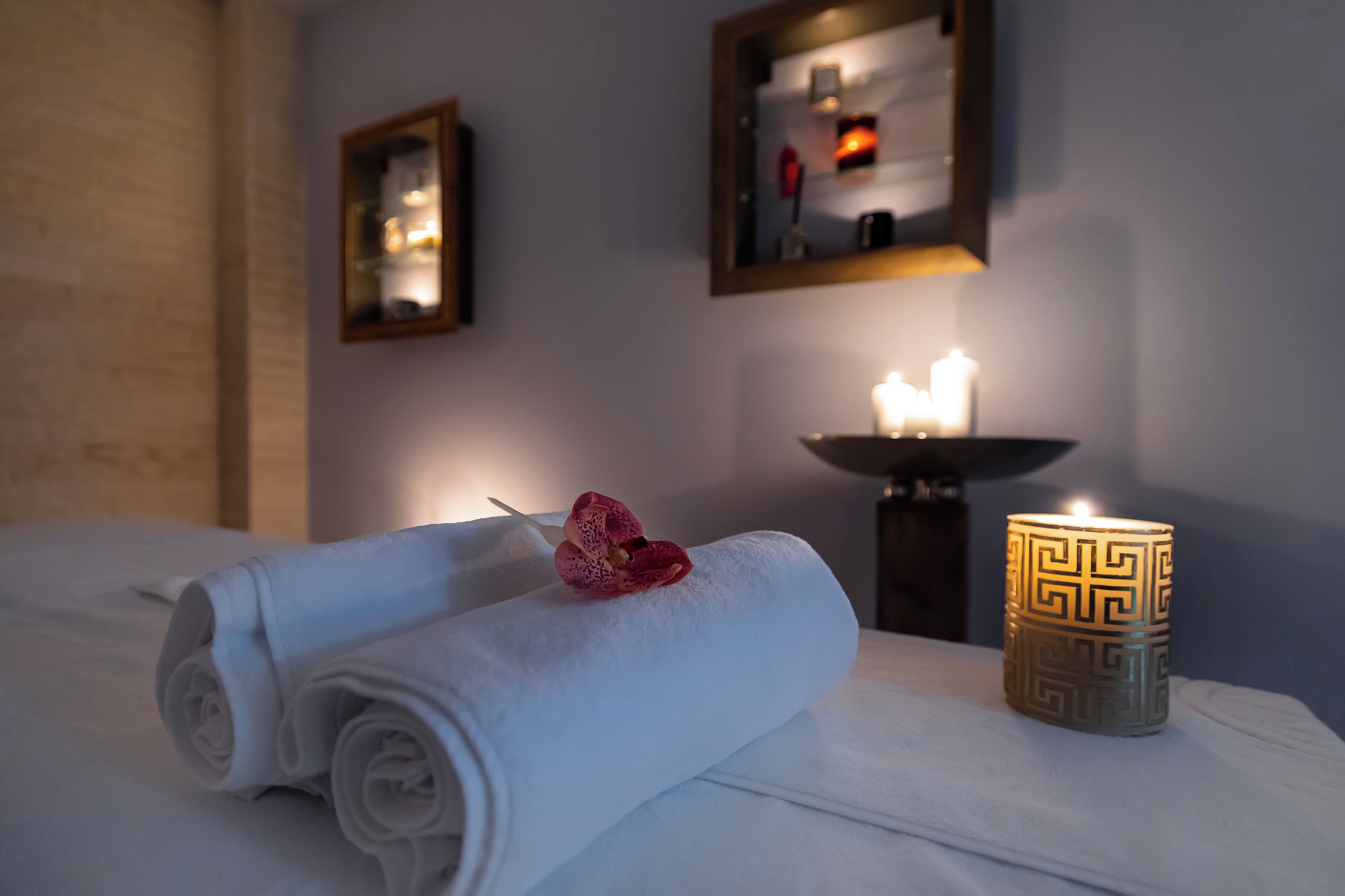 A neatly made bed with rolled white towels and a decorative pink orchid, a lit candle in a patterned holder, and a mirror on the wall.