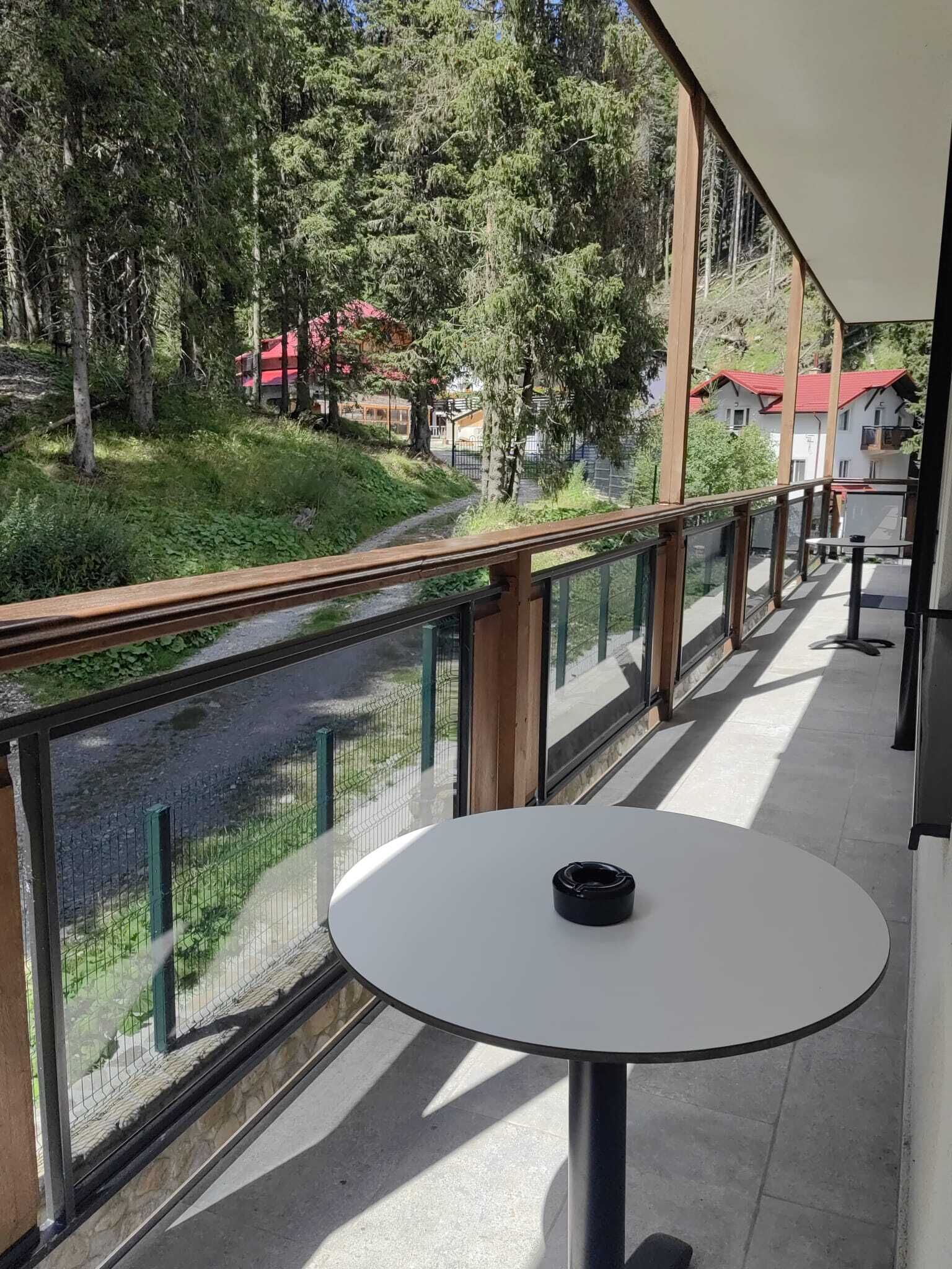 En balkon med et bord og stole, der ud til en flod og et skovområde med bygninger i baggrunden.