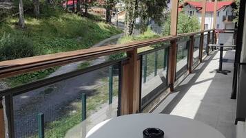 En balkon med et bord og stole, der ud til en flod og et skovområde med bygninger i baggrunden.